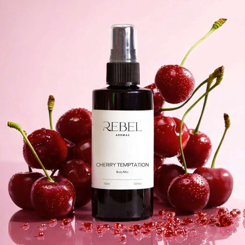 Bottle of Rebel Aromas Cherry Temptation Body Mist surrounded by cherries on a pink background