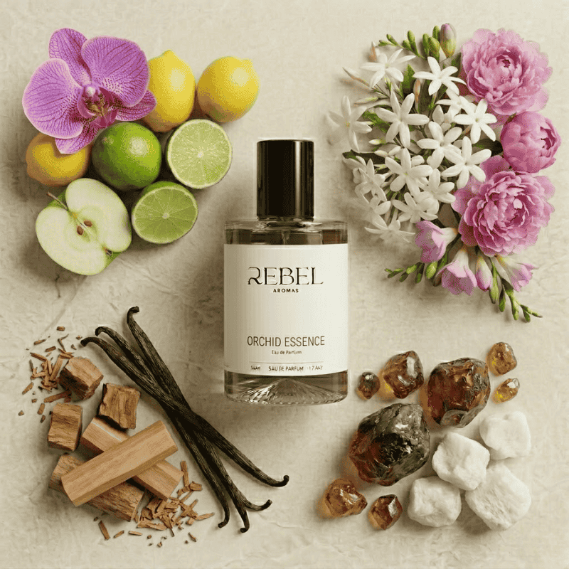 Rebel Aromas Orchid Essence perfume bottle surrounded by flowers, fruits, and vanilla beans on a textured surface.