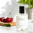 REBEL Aromas Opium Red perfume bottle on a table with cherries and a plant in the background

